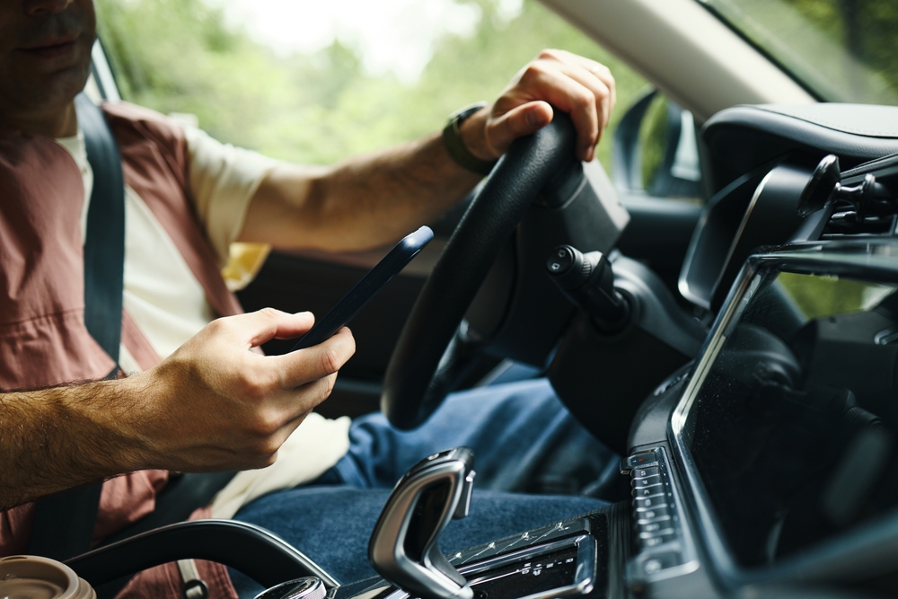 Caucasian young adult man driving car while holding smartphone in one hand, sitting in driver seat during family road trip, dashboard and steering wheel visible in foreground